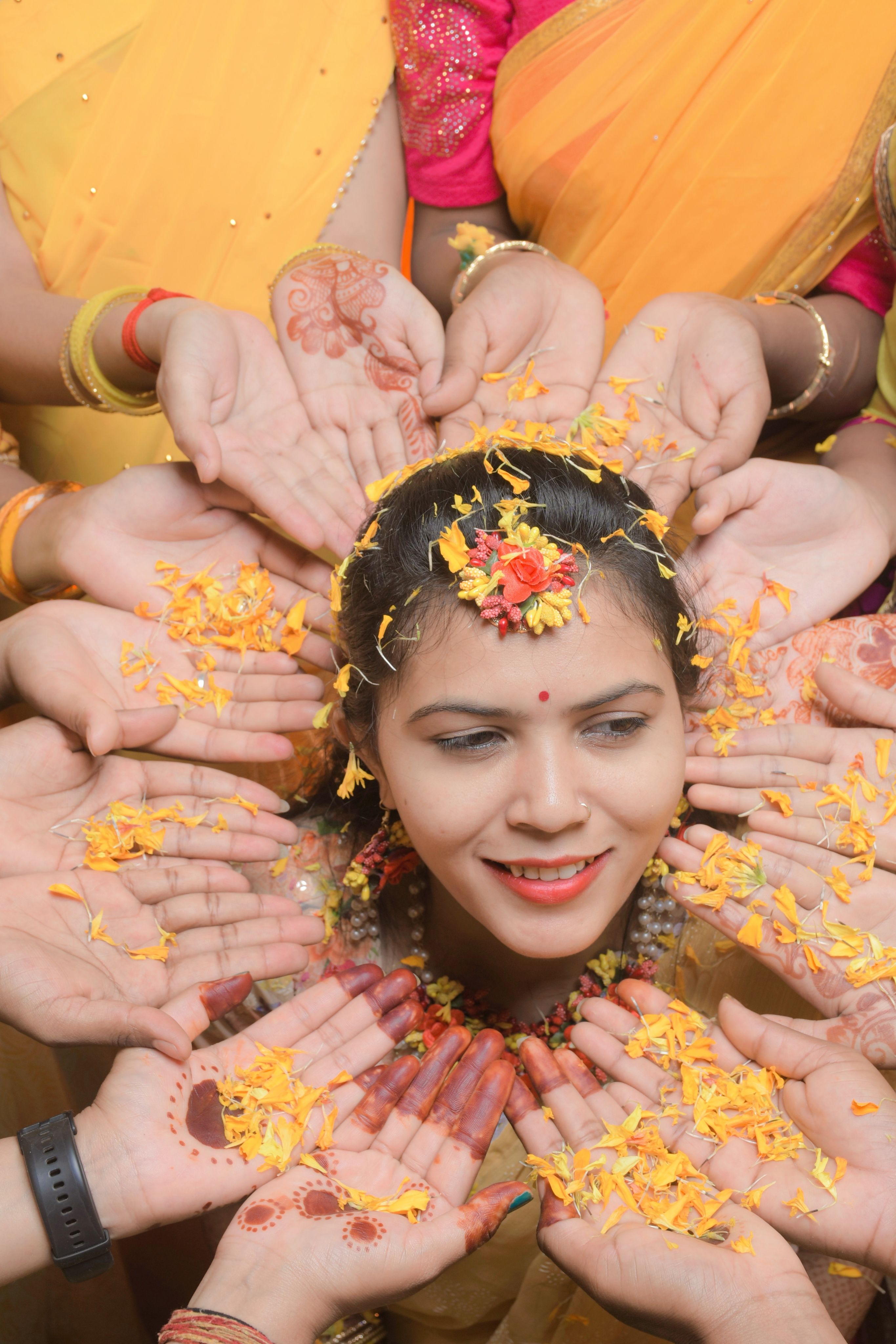 Smiling Indian bride surrounded by supportive hands and flower petals, showcasing traditional celebration.