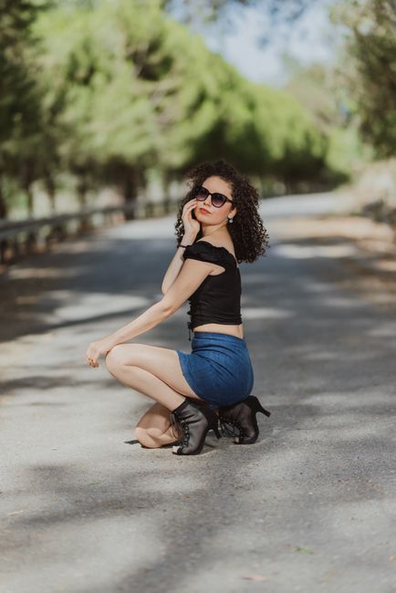 Fashionable woman in sunglasses posing on a sunny road, showcasing modern style with blouse and skirt.