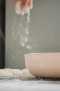 Close-up of a hand sprinkling flour into a bowl beside dough, showcasing baking preparation.