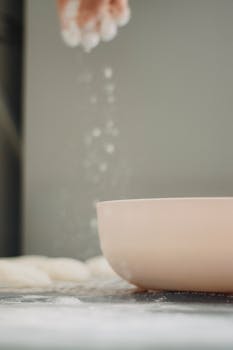 Close-up of dough preparation with flour sprinkling into bowl, creating indoor baking scene.