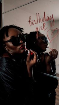 Chic woman in sunglasses applying lipstick, with 'birthday girl' written on the mirror.