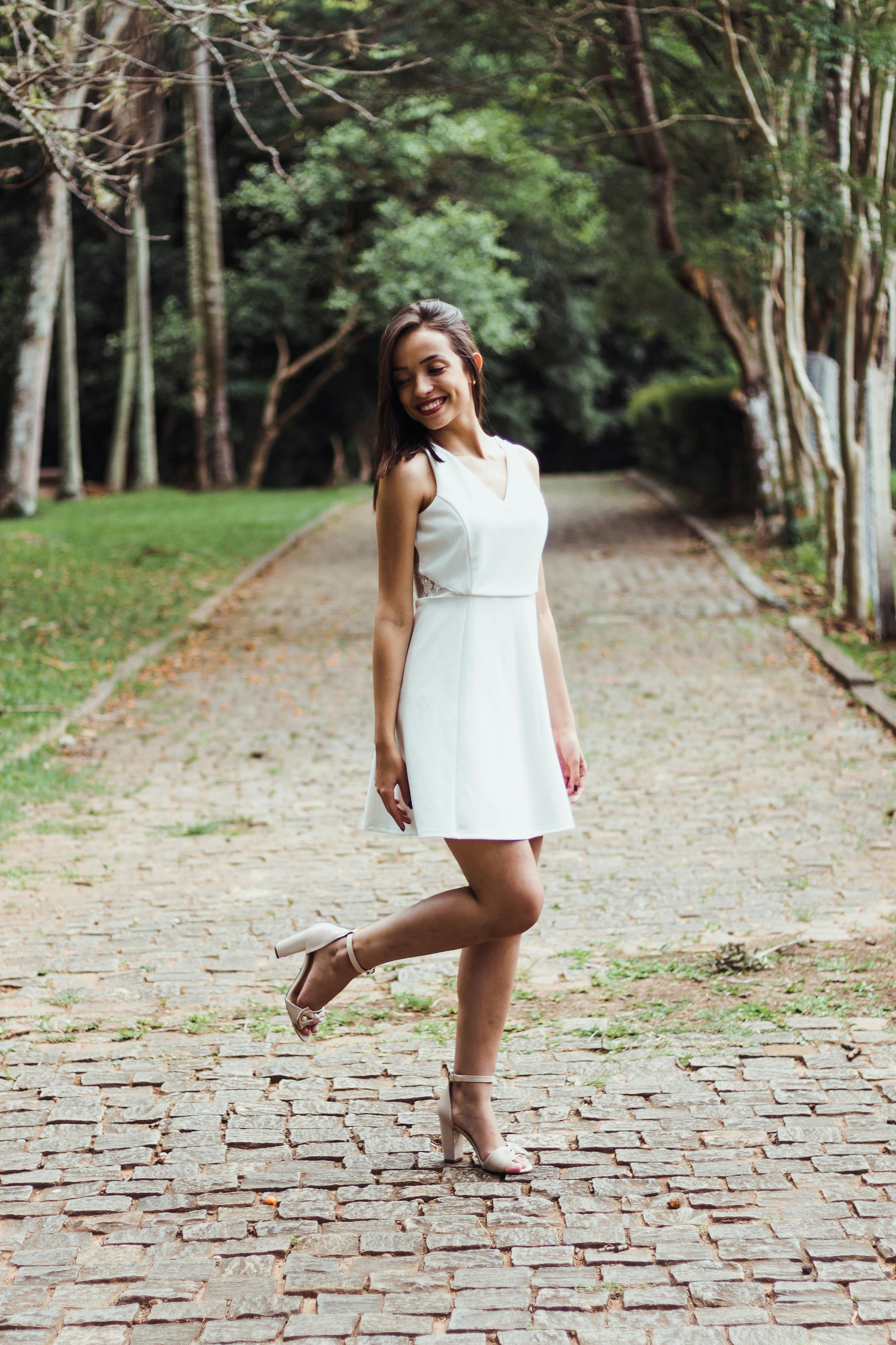 一位身穿白色连衣裙的时尚女子，在绿树环绕的户外鹅卵石小路上摆着姿势