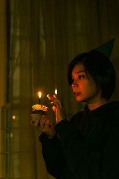 A young woman with a party hat holds a cupcake with a lit candle in a dimly lit room, creating a warm and intimate atmosphere.