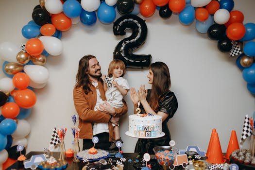 A family celebrating a toddler's second birthday with cake and colorful balloons.