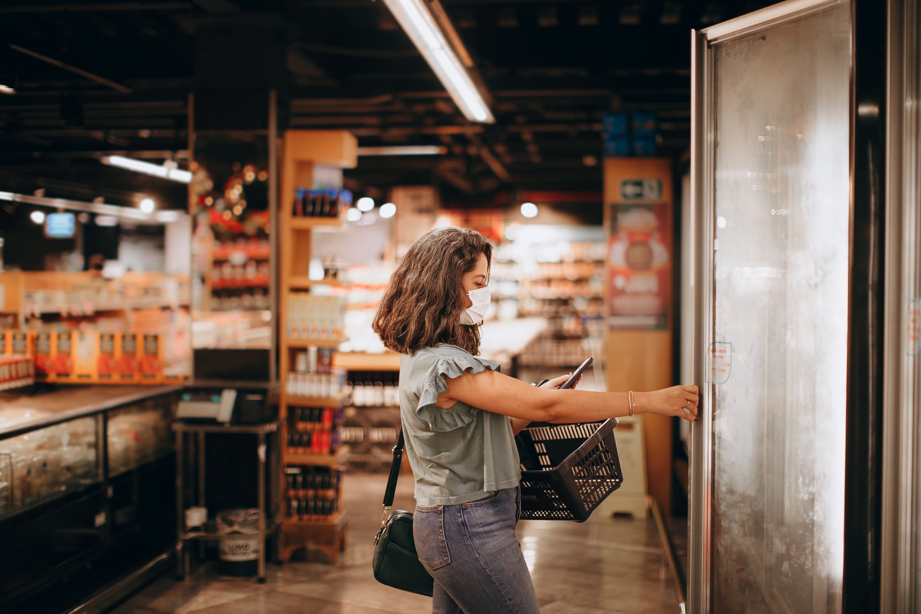 Woman wearing mask shops in grocery store, reaching for freezer door, holding basket.