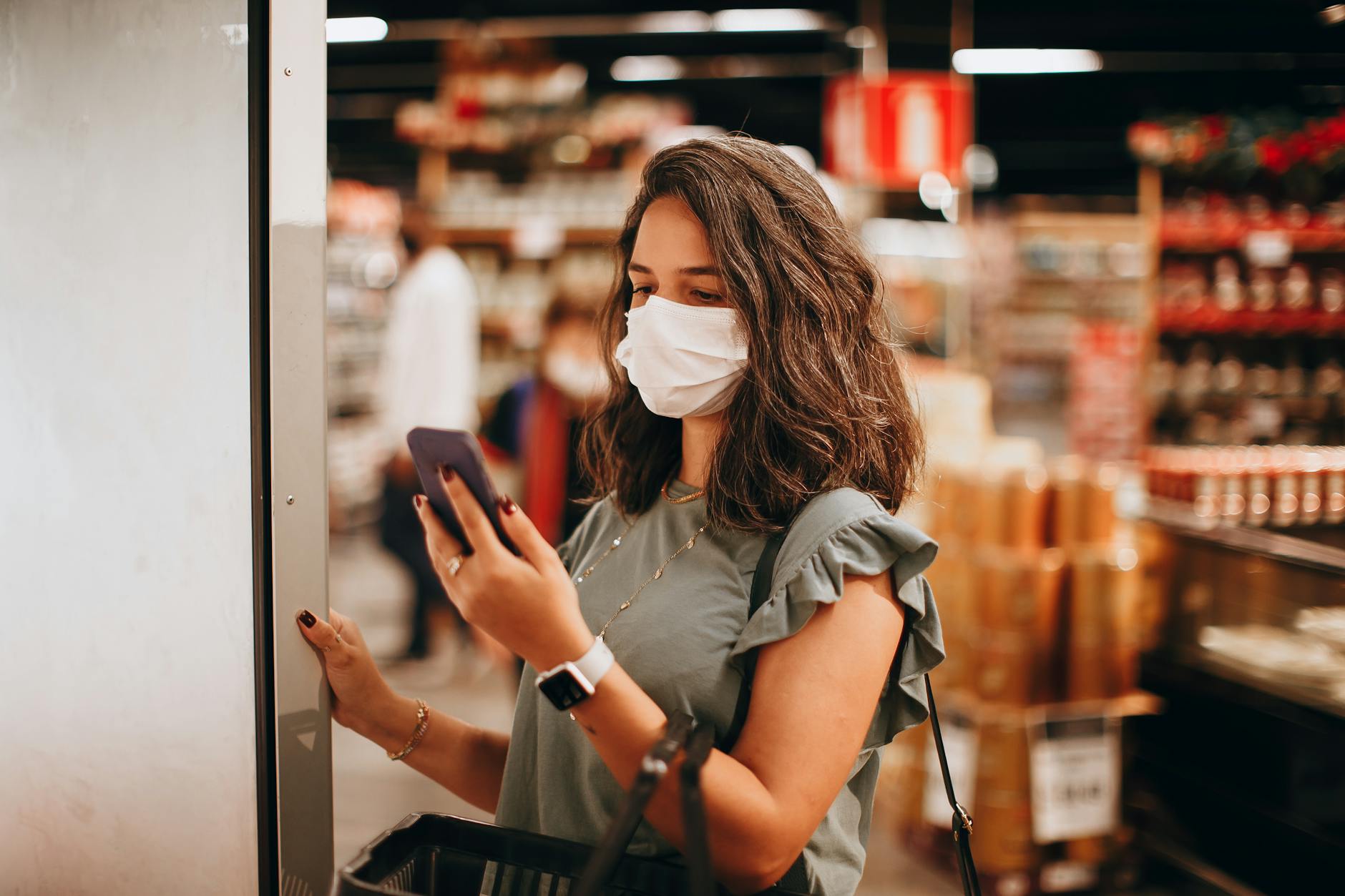 Woman wearing mask shopping in a supermarket, checking smartphone. Captures modern retail experience.
