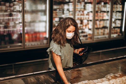 A woman in a mask selects items from a supermarket freezer aisle with a shopping basket.