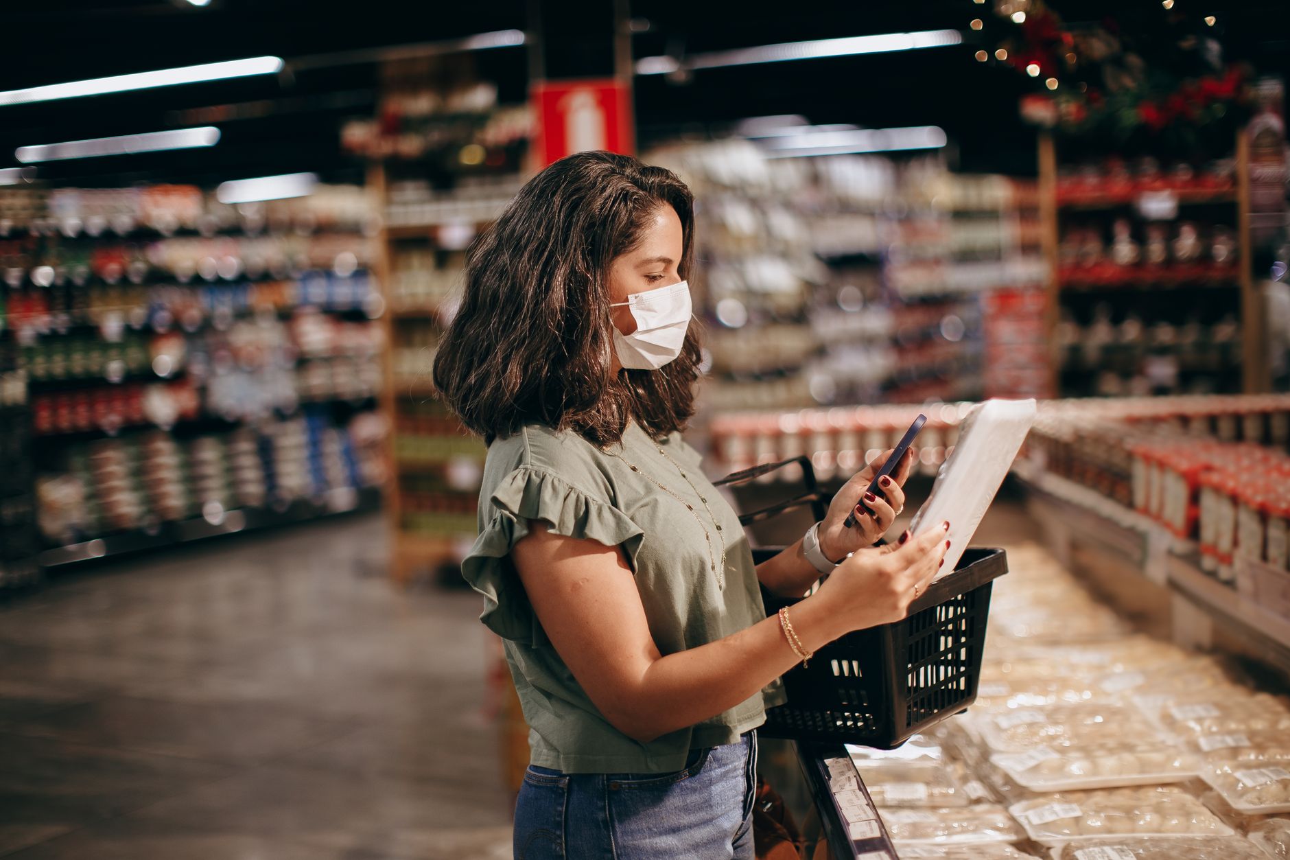 A woman shopping in a supermarket, wearing a face mask and using her phone to check products.