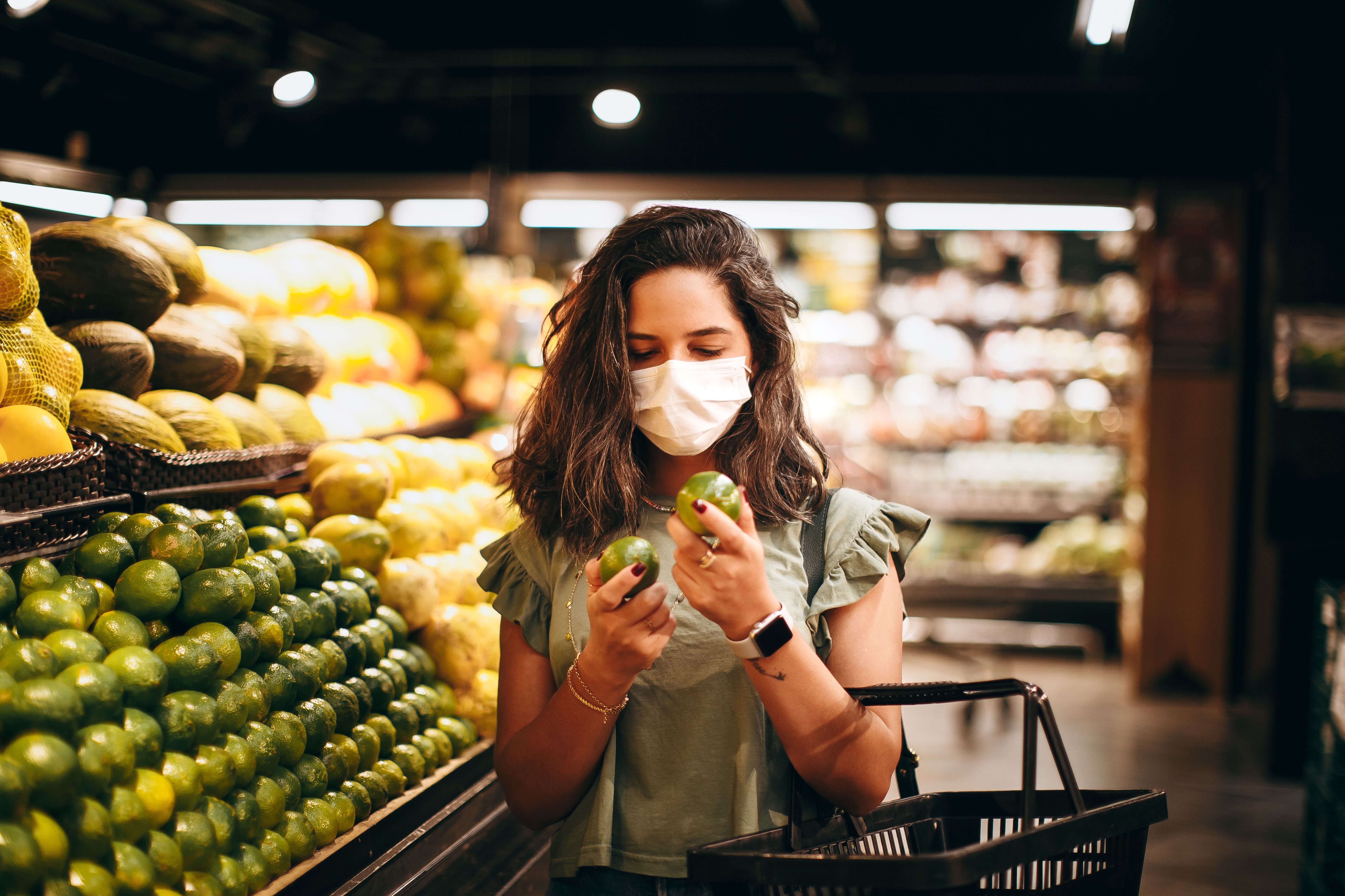A woman wearing a mask shops for green apples in a supermarket produce section.
