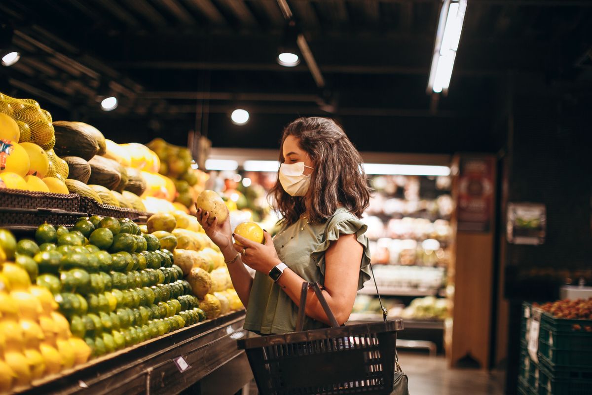 Woman wearing a mask choosing fruits in a supermarket aisle, focusing on a safe shopping experience. Illustrative editorial photograph.