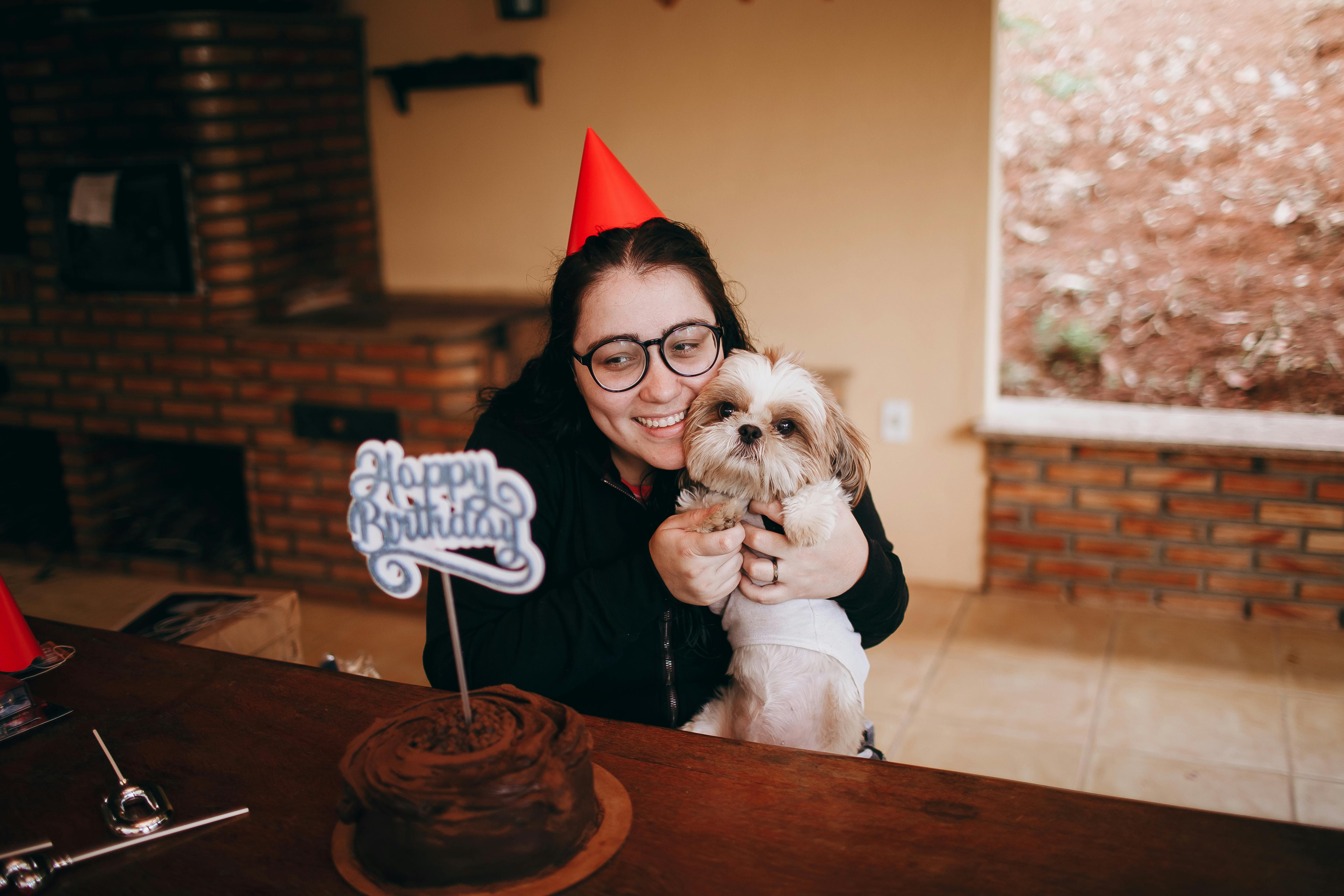 A joyful birthday celebration indoors with a woman hugging her dog by a cake.
