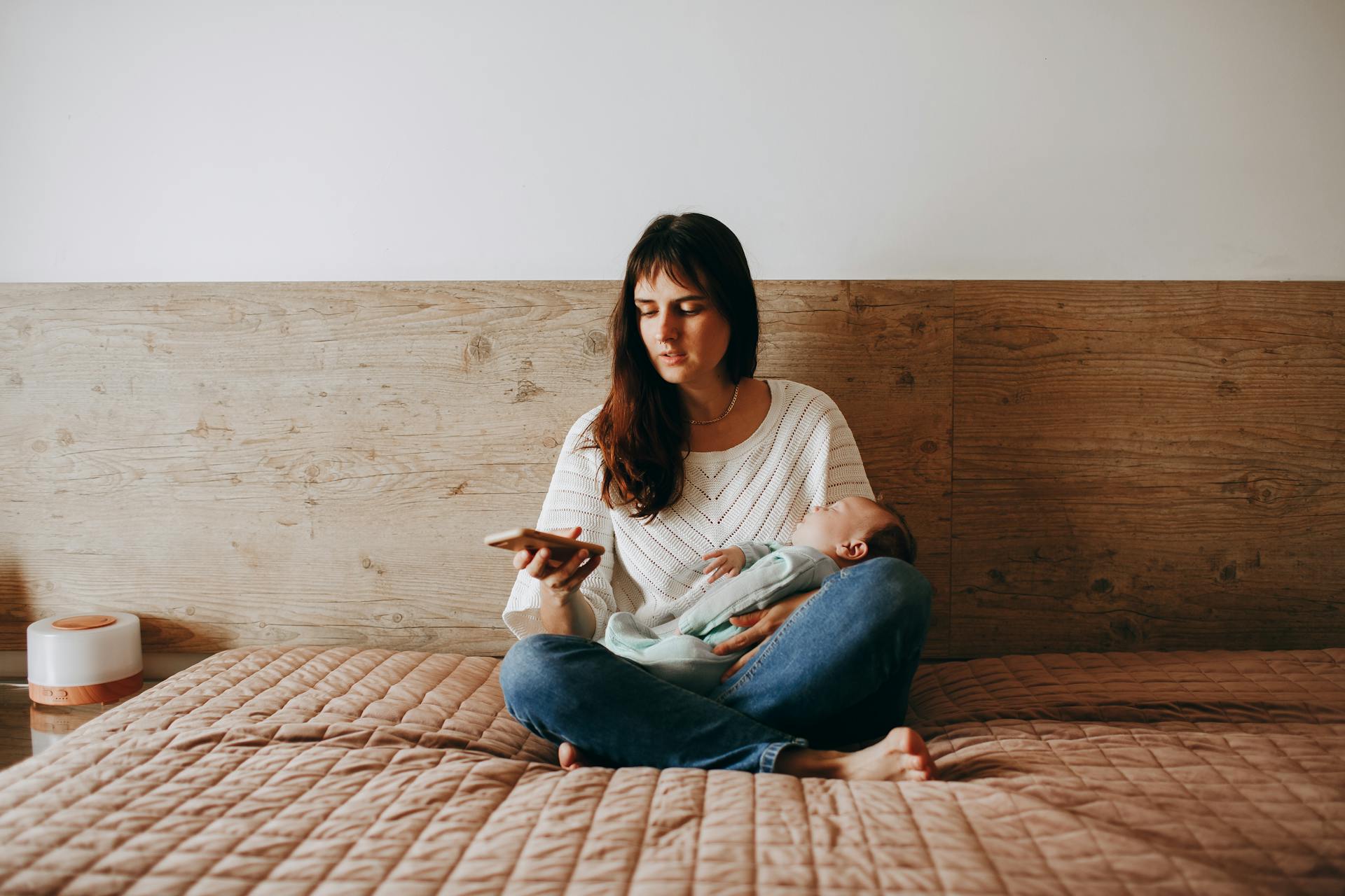 A woman sitting on a bed holding a baby and using a cell phone