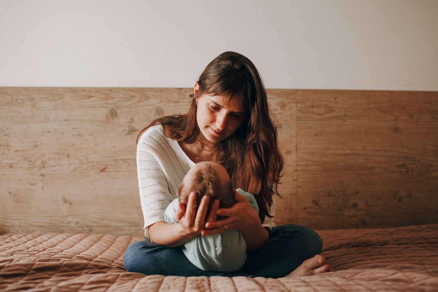Mother seated indoors holding her baby with a simple wood headboard behind them