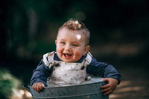 Free Charming baby with curly hair smiling happily while sitting in a tin bucket outdoors. Stock Photo