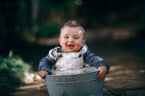 Free Smiling baby sitting in a metal tub outdoors, wearing overalls and a long-sleeve shirt. Stock Photo