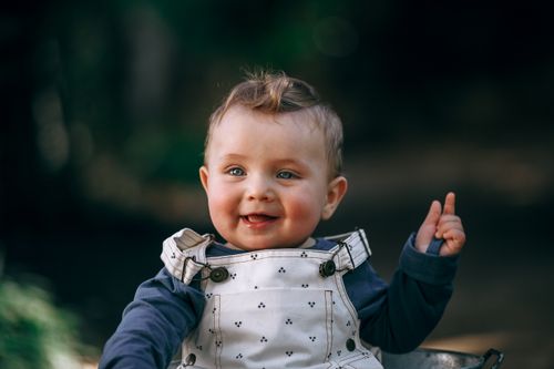 Free Charming baby in overalls smiles brightly against an outdoor blurred background. Stock Photo