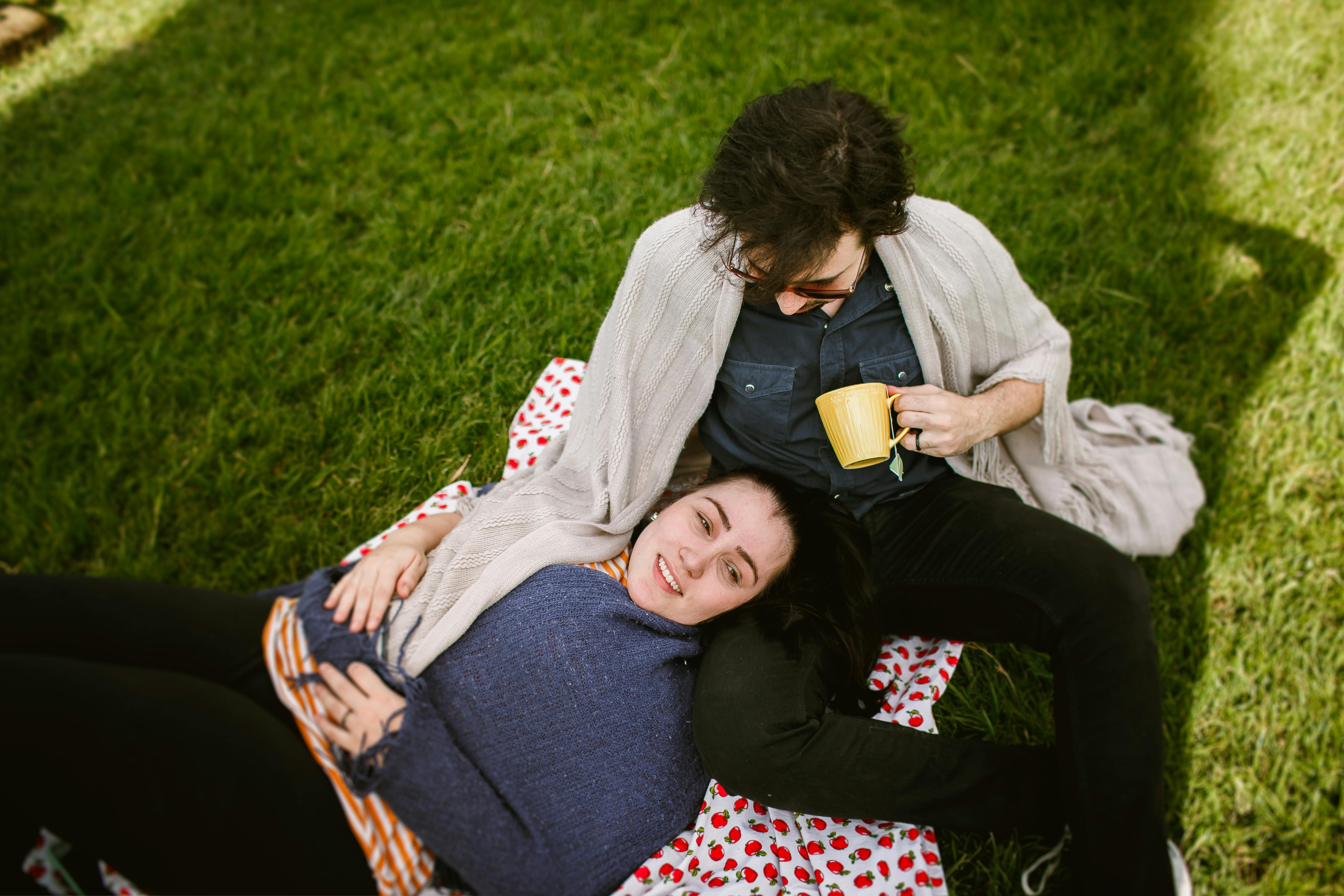 A happy couple enjoying a sunny day outdoors on a blanket with a mug in hand.