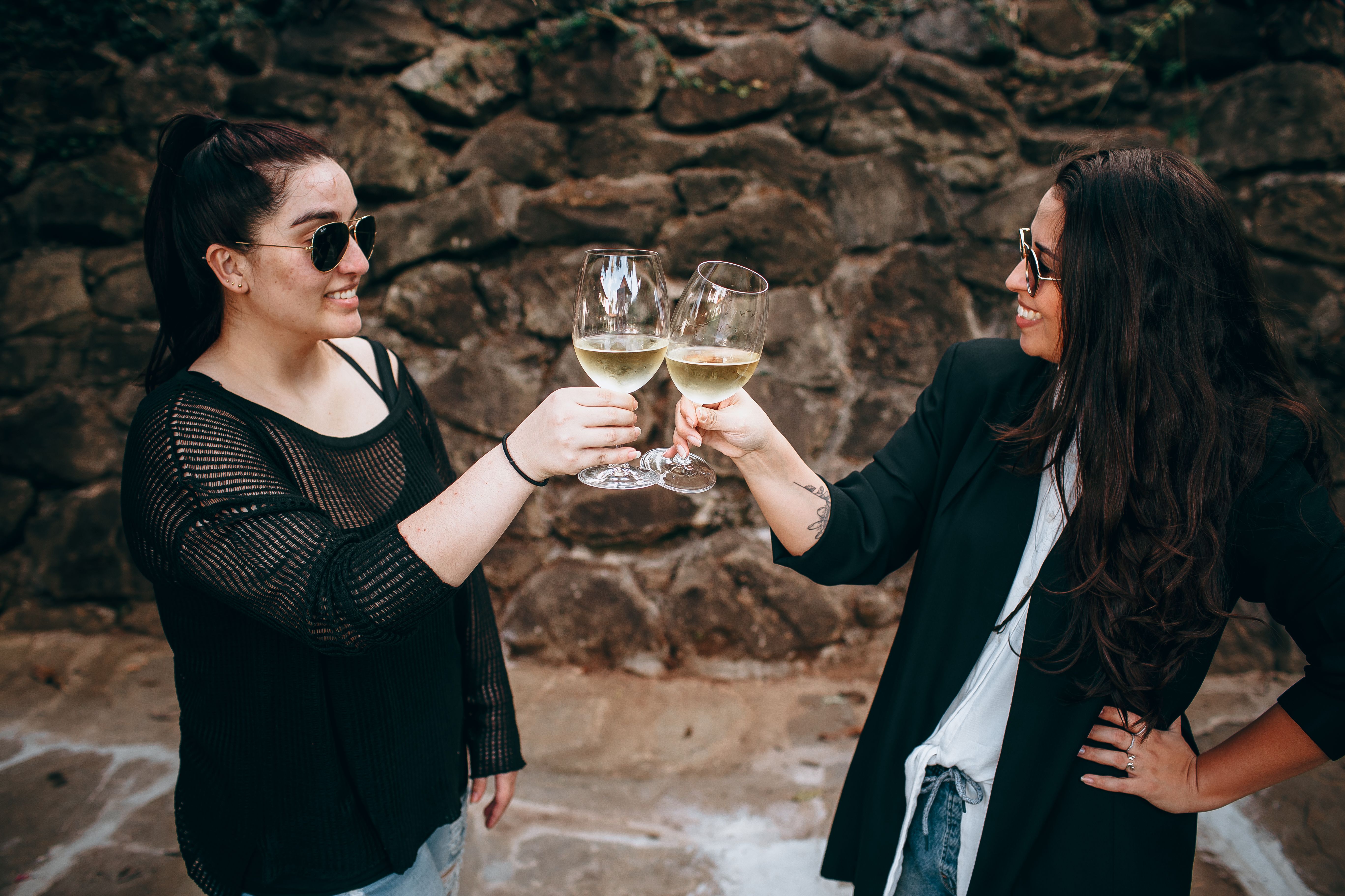 Two women in casual attire enjoying an outdoor wine toast on a stone backdrop.