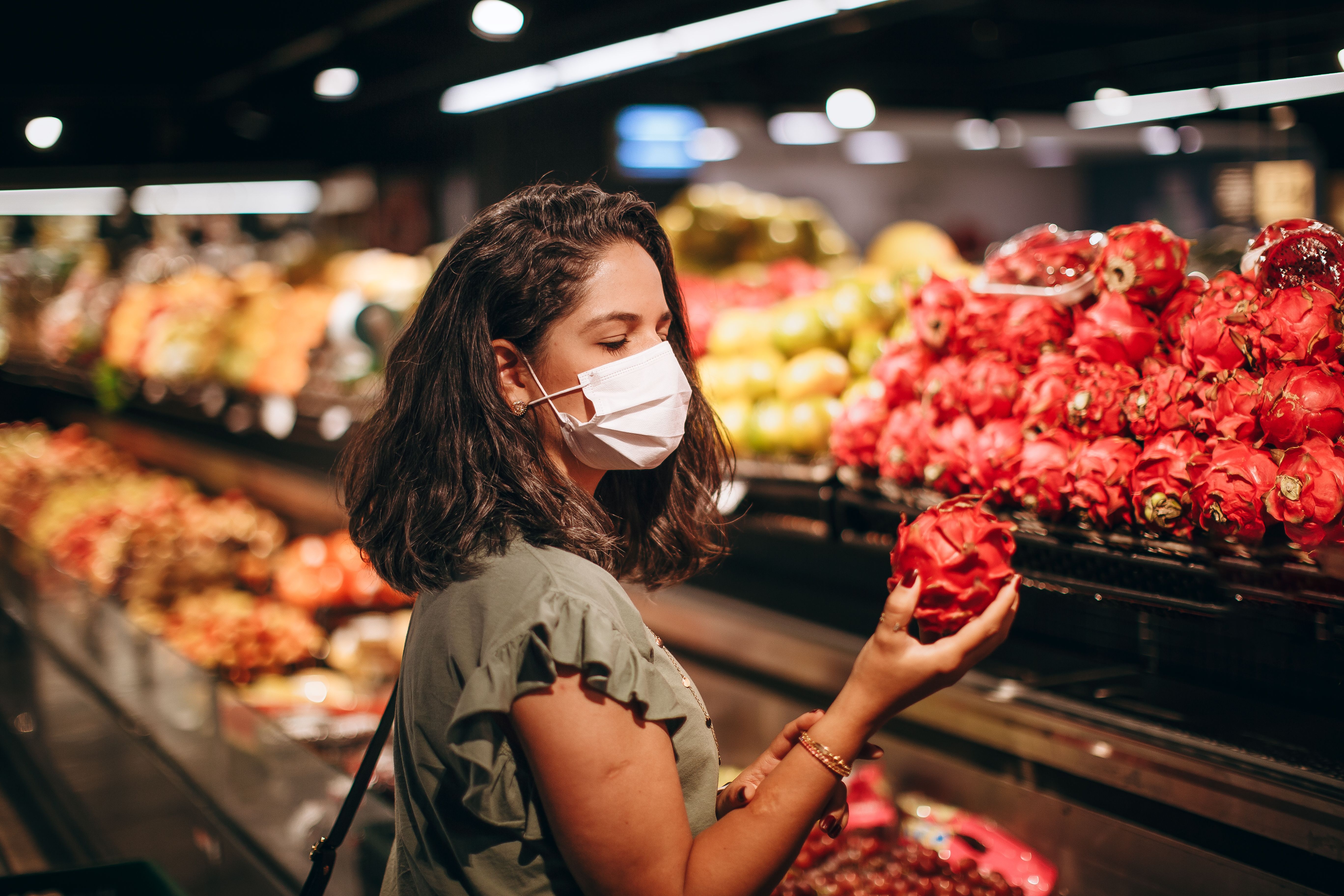 Woman wearing a mask while selecting fresh dragon fruit at a supermarket fruit aisle.