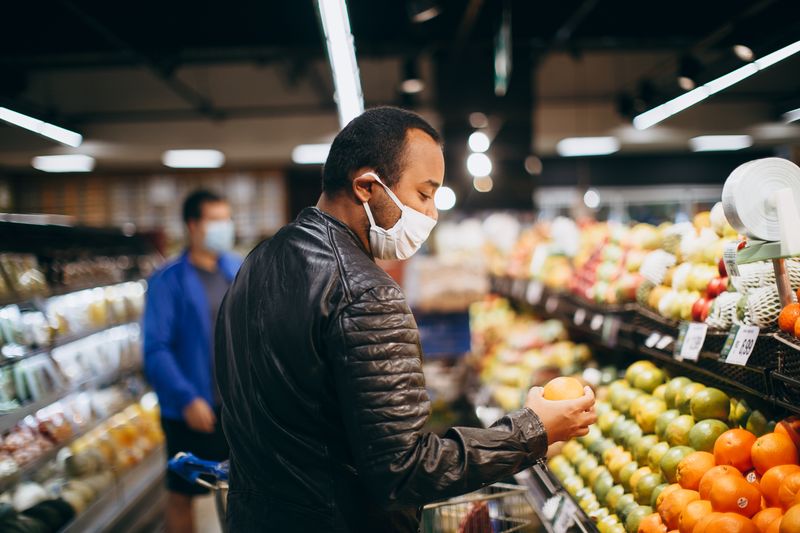 Man wearing mask selecting fresh fruit in a supermarket aisle.