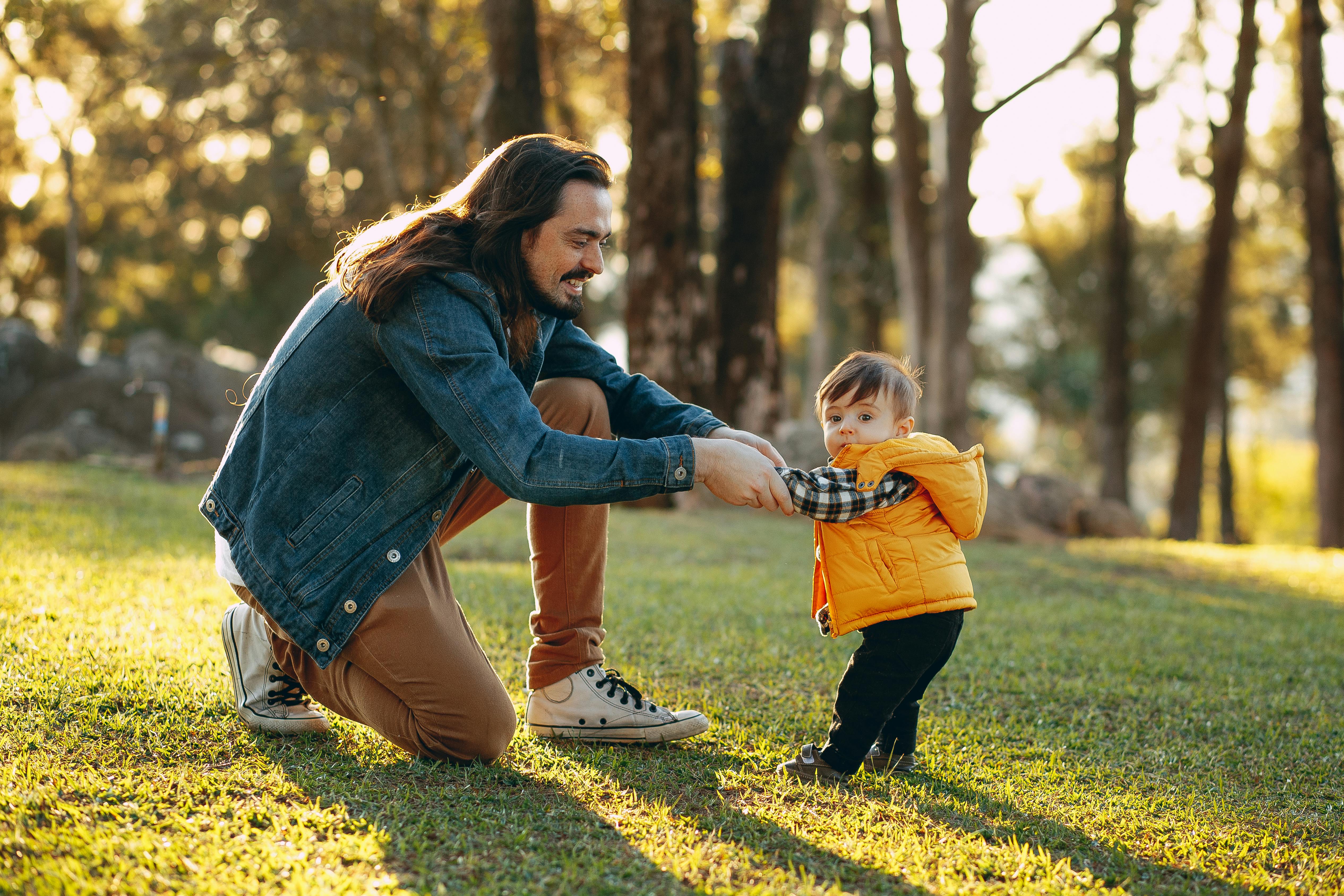 A father playing and holding the hands of his toddler in a sunlit outdoor park setting.