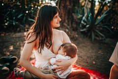A serene scene of a mother breastfeeding her baby while sitting on a blanket outdoors in summer.