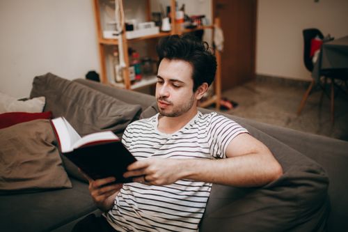 Free Man in a striped shirt relaxing on a sofa, reading a book indoors. Stock Photo