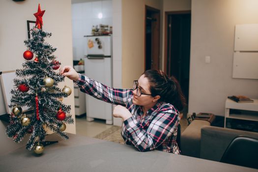 A woman decorates a small Christmas tree with ornaments in a cozy living room.
