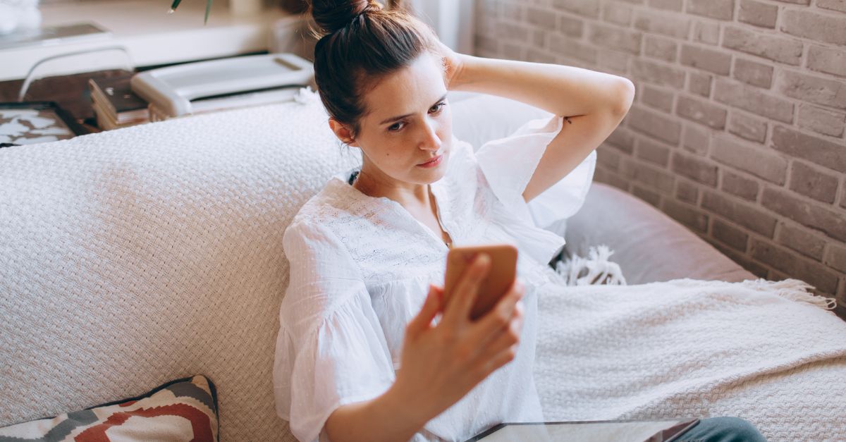 Uma mulher ajusta o cabelo enquanto tira um selfie com um smartphone, sentada em um sofá com um tablet, personificando o estilo de vida do trabalho remoto.