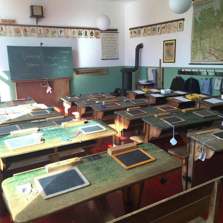 An old-fashioned schoolroom in which we look from the back of the classroom over all the slates arranged at each student's desk.