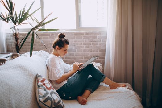 A woman comfortably seated on a sofa, working remotely using a tablet, in a stylish living room setting.