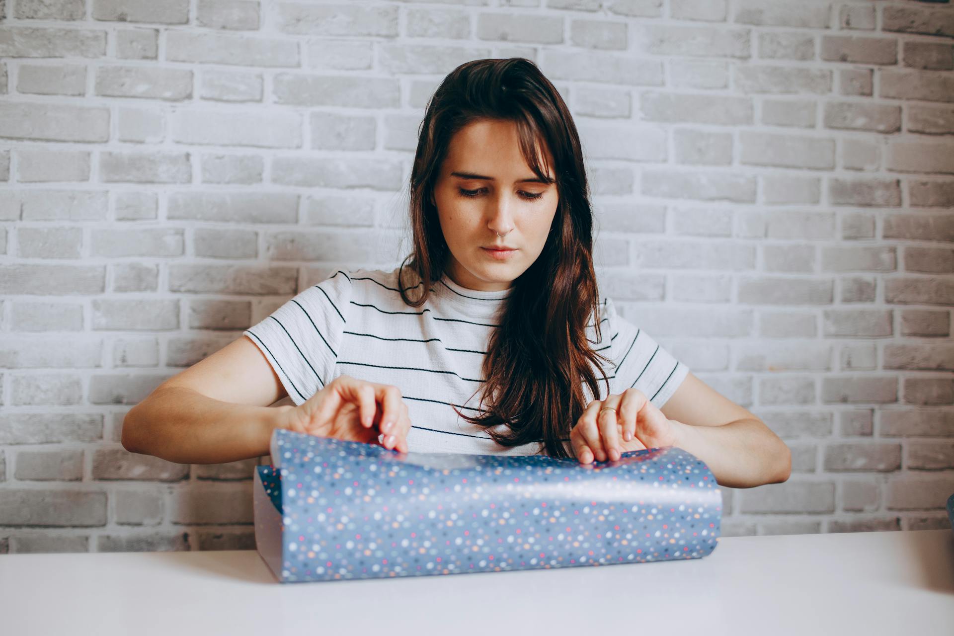 Young woman focused on wrapping a gift against a stylish brick wall indoors.