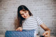 A young woman with brunette hair wrapping a gift in blue paper against a brick wall.