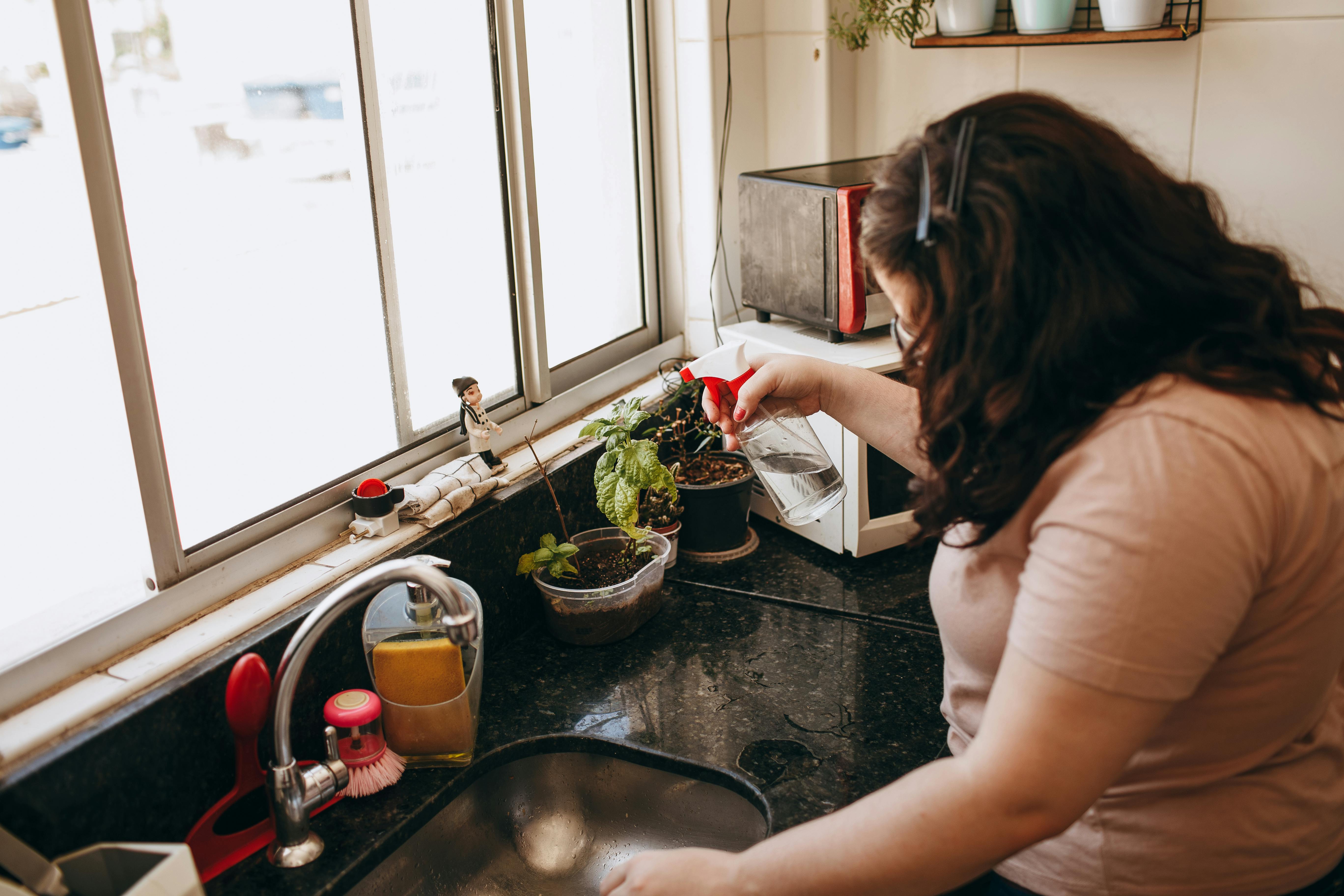 A woman cares for indoor plants by the kitchen window using a spray bottle.