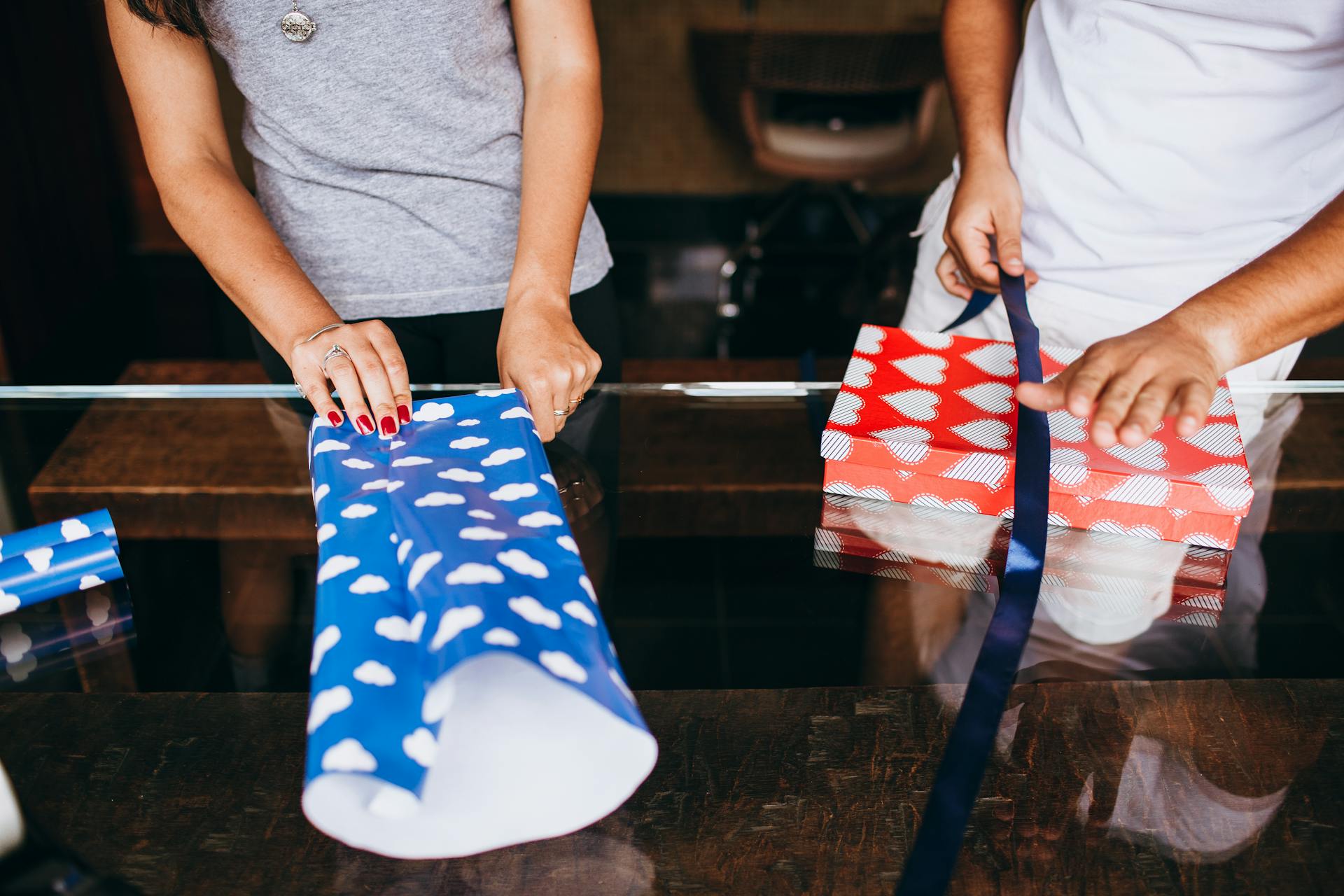 Two adults wrapping gifts with colorful paper and ribbons indoors.