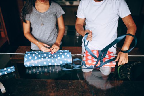 Free A man and woman wrapping gifts with cloud-patterned paper and ribbon on a glass table. Stock Photo