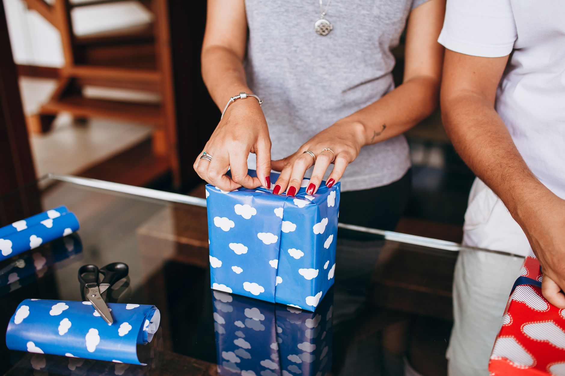 Two adults wrapping gifts with blue cloud-patterned paper indoors on a glass table.