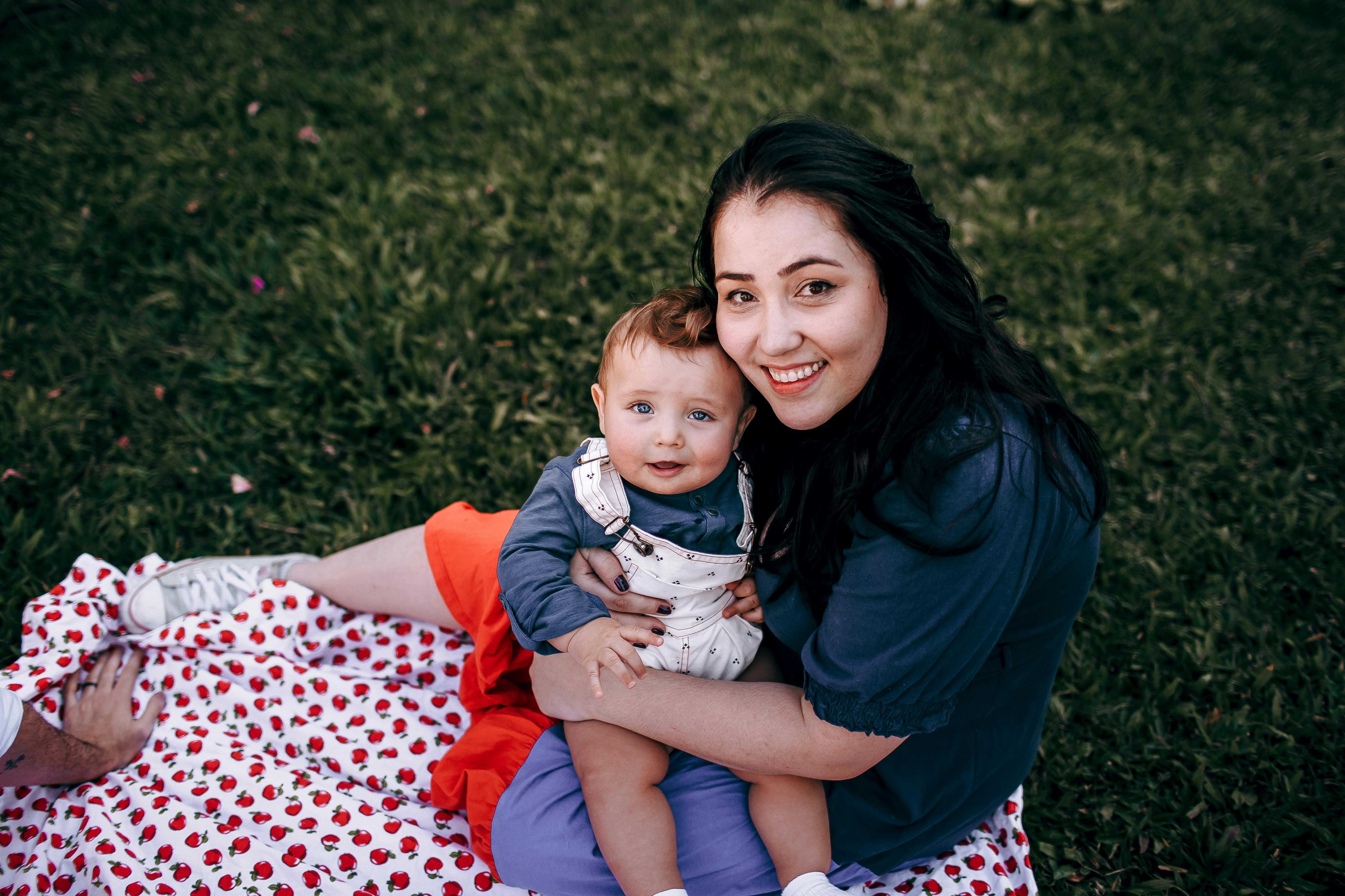 A mother and her baby enjoy quality time outdoors on a picnic blanket surrounded by greenery.