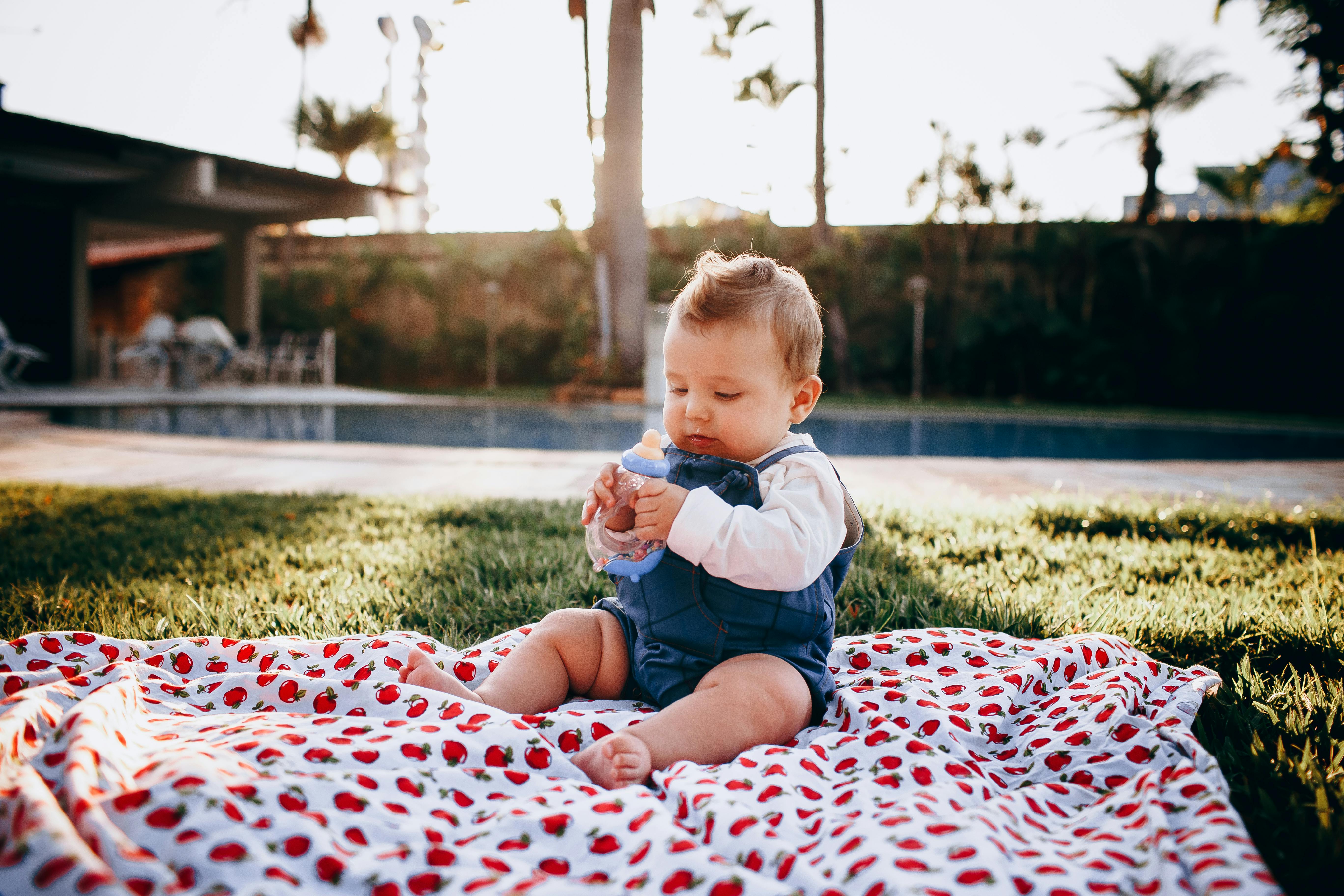 A cute baby sitting on a blanket in a sunny backyard, holding a bottle and enjoying a picnic.