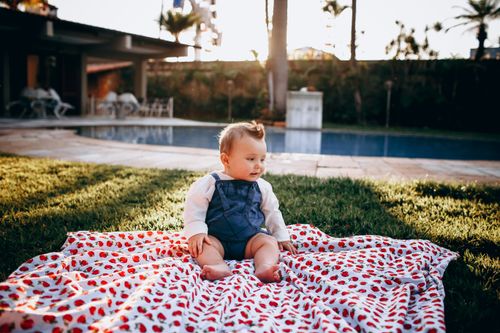Free Adorable baby enjoying a sunny day on a strawberry-patterned blanket by a pool. Stock Photo