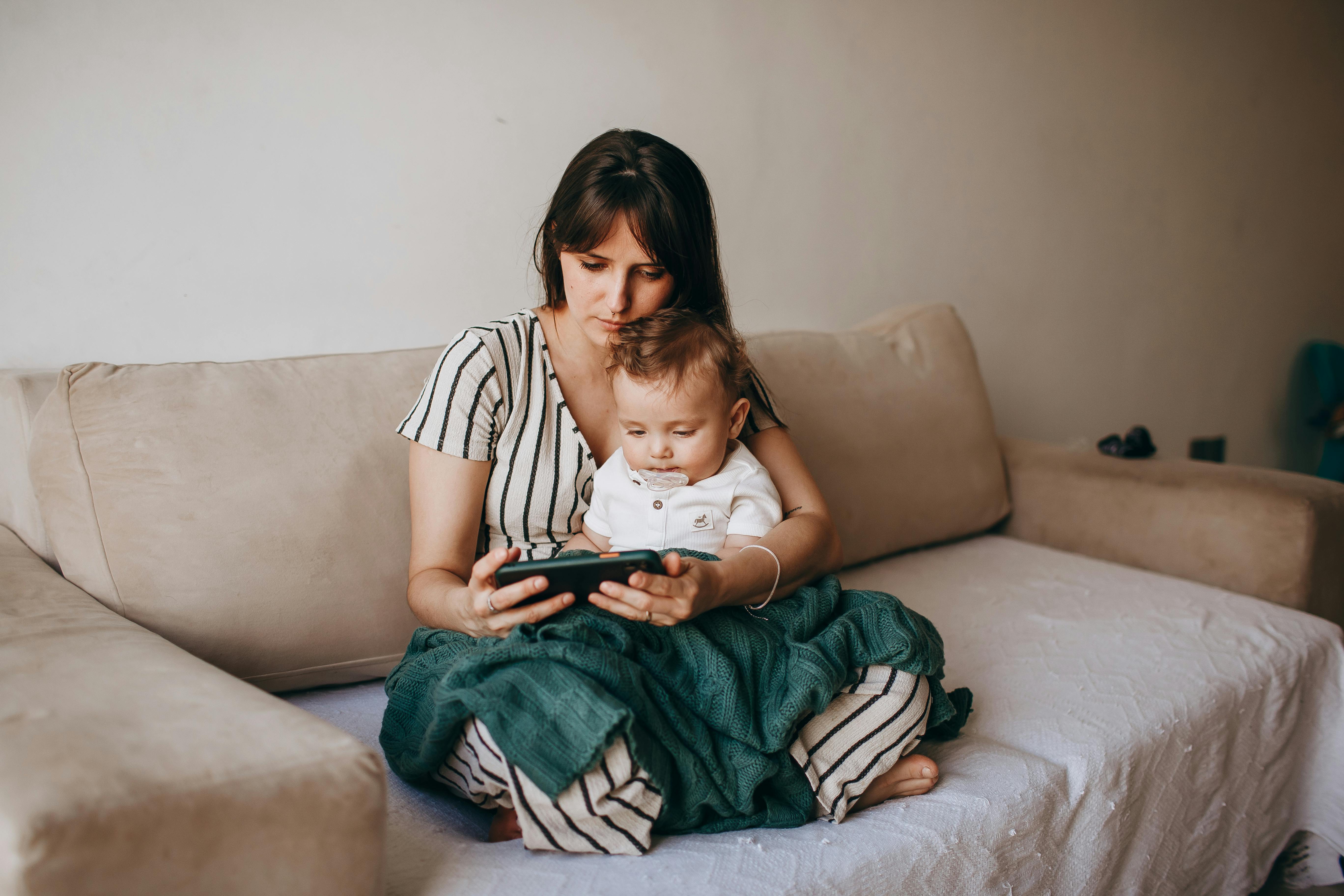 A woman and her baby sit together on a beige sofa, engaging with a tablet device.