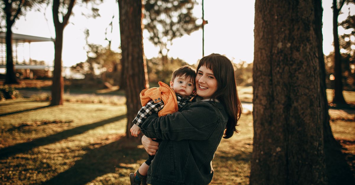 Smiling mother holding her baby in a sunlit park, surrounded by trees.