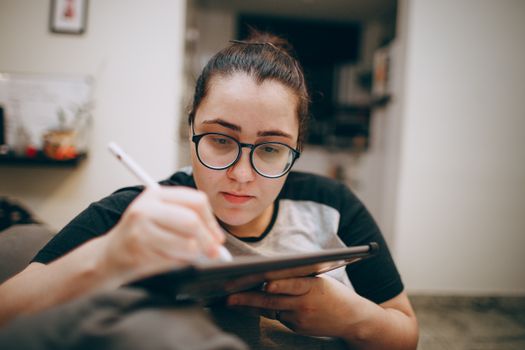 Young woman wearing glasses, concentrating while writing on a digital tablet indoors.