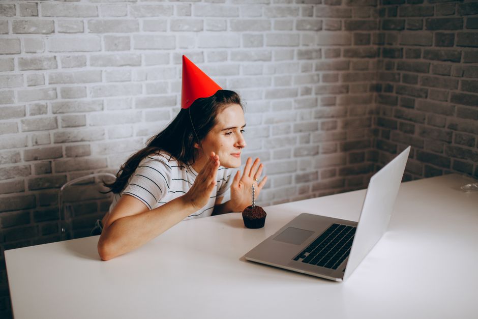 Woman in party hat on video call celebrating birthday with cupcake.