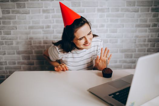 Woman in party hat waving at laptop and cupcake, celebrating virtual birthday.