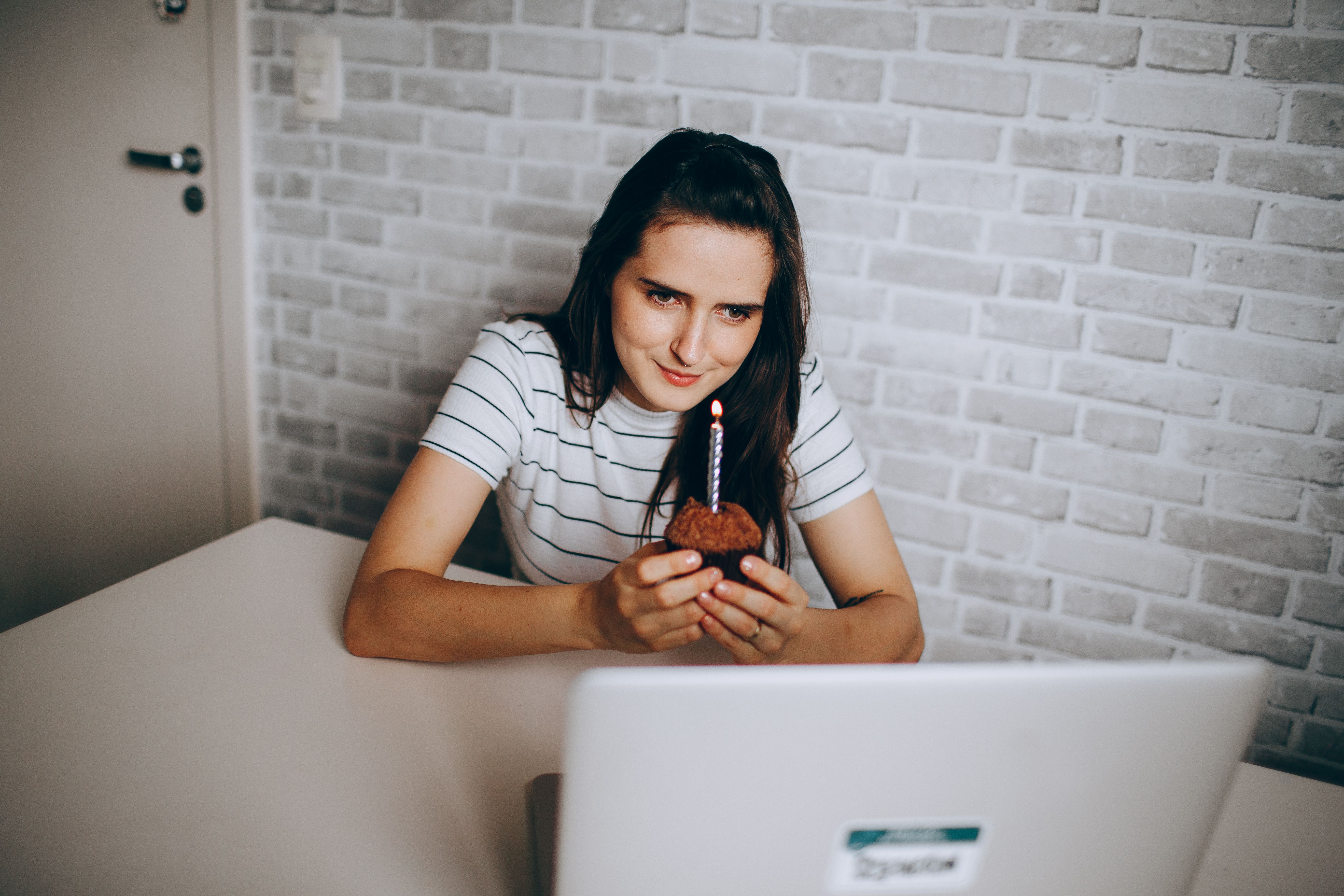 Woman enjoying a birthday celebration with a cupcake and lit candle during an online video call.