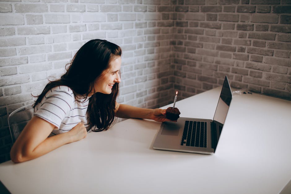 Woman smiling at laptop during a birthday video call, holding a cupcake with a lit candle.