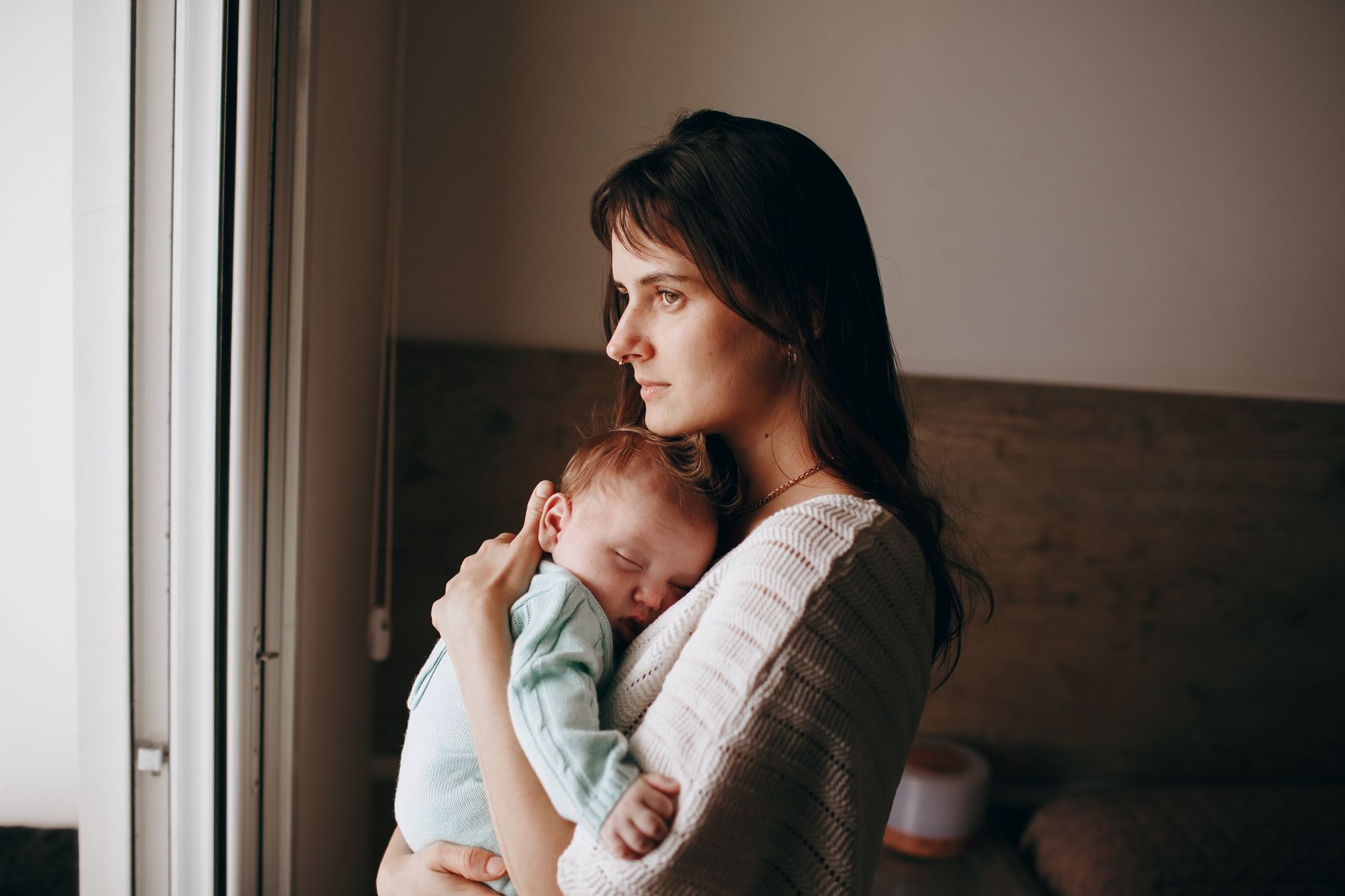 A serene moment of a mother holding her sleeping baby near a window, indoors.