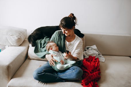 A woman lovingly holds her baby on a cozy sofa with a dog nearby, indoors.