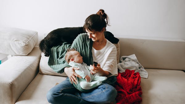 A woman lovingly holds her baby on a cozy sofa with a dog nearby, indoors.